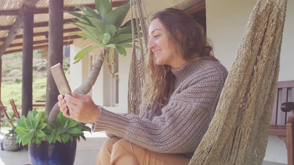 Smiling caucasian woman lying in hammock using tablet on rural garden terrace