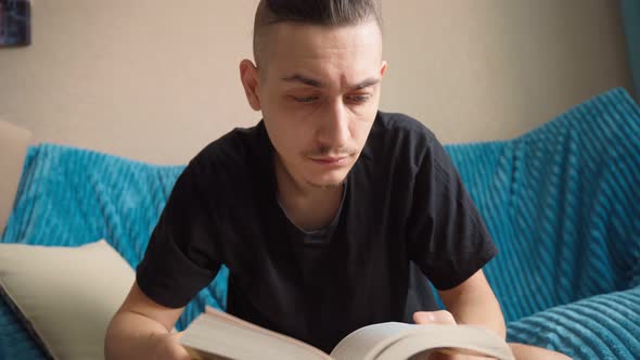 Man Thoughtfully Reading Book is Sitting on the Sofa Wide Angle