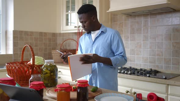 Focused Young Man Writing with Pen Examining Ripe Harvested Vegetables in Kitchen