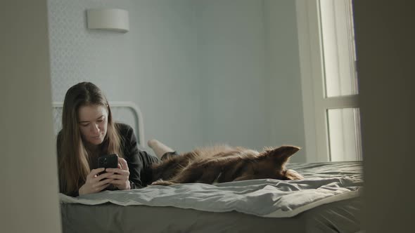 Young Woman At Home Using Her Smartphone Close To Her Dog.