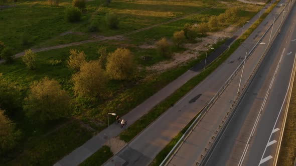 Aerial Backside View: Two People Ride Bicycles Near Highway Bridge During Sunset