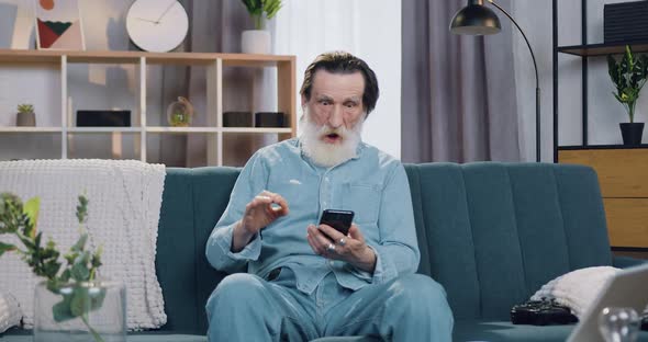 Bearded Man Got Good News on Smartphone and Rejoycing with Raised Hands Sitting on the Couch at Home