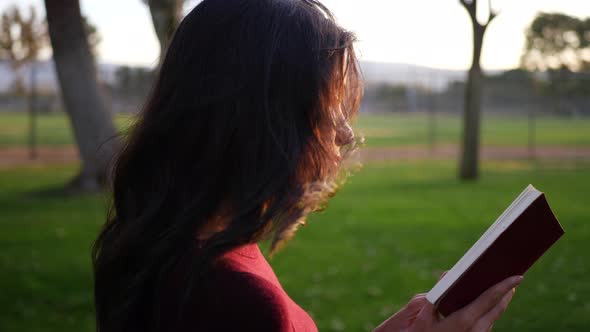 A beautiful young woman reading the pages of a red book outdoors in the park at sunset SLOW MOTION.