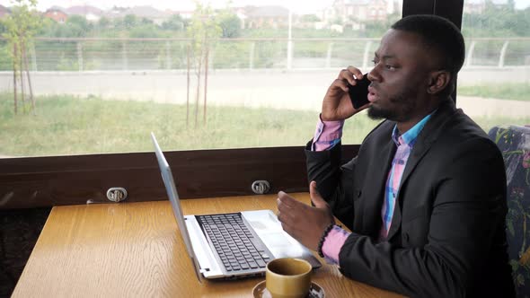 Black Businessman Works on Laptop and Calls Smartphone Sits in Cafe with Coffee