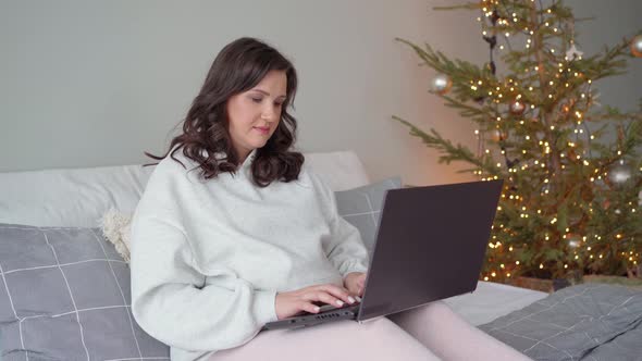 Beautiful Young Pregnant Woman in the Bedroom Sits on the Bed Next to the Christmas Tree and Works