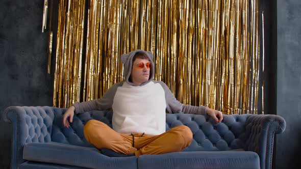Young Man in Hood with Ears and Bright Glasses Sitting on Sofa Against Wall with Golden Tinsel