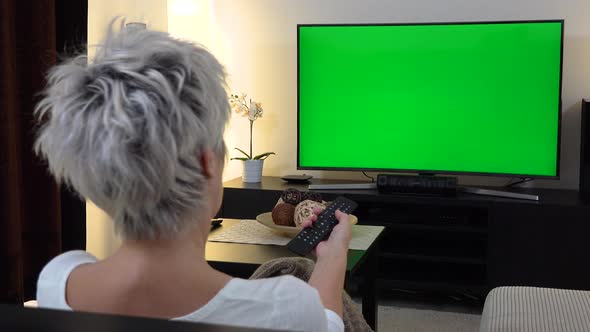 A Middle-aged Woman Sits, Watches TV with a Green Screen and Switches Programs with a Remote Control