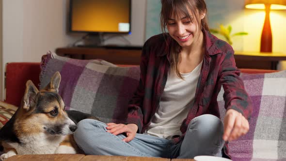 Tender Woman Sits on Couch with Dog Hugging and Feeding Him