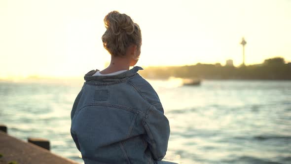 Back View of Young Pretty Blond Girl Sitting on Concrete Pier Enjoying Warm Sunset Shine Looking at