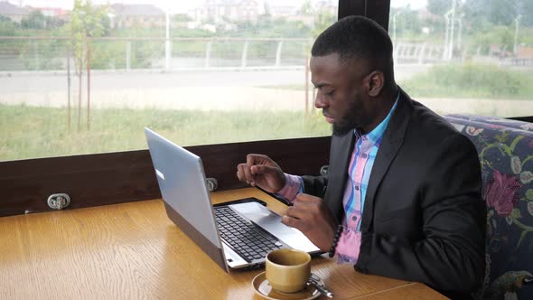 Black Businessman is Working Typing a Message on Laptop Sitting in Summer Cafe