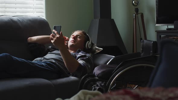 Relaxed caucasian disabled man lying on sofa in living room wearing headphones and using smartphone