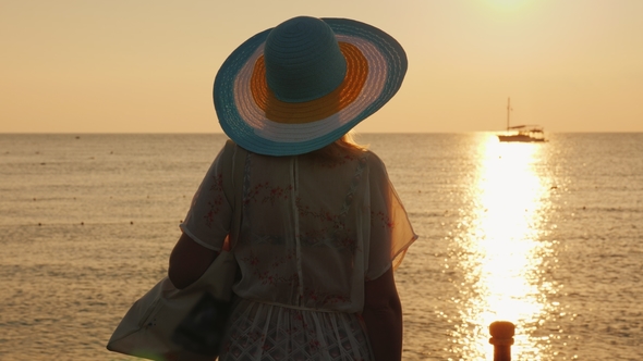 A Young Woman in Beach Clothes and a Wide-brimmed Hat Is Enjoying the Sunrise on the Seashore