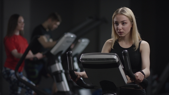 Nice Girl in Blak Sport Wear Vigorously Works on Exercise Bike and Watchs Time with Her Wrist Watch