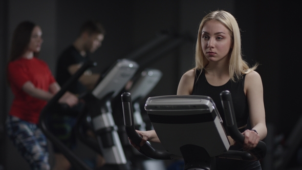 Cute Girl in Blak Sport Wear Vigorously Works on Exercise Bike and Watchs Time with Her Wrist Watch