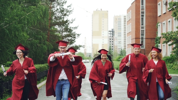 Excited Graduating Students Running Along Road on Campus Holding Diplomas Wearing Graduation Clothes