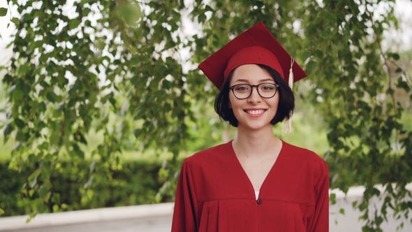 Portrait of Joyful Young Woman Graduating Student in Gown and Mortar-board Smiling and Looking at