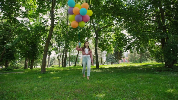 Happy Young Girl Walks in the Green Sunny Park and Holds Colourful Balloons