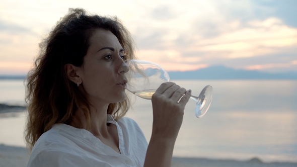 A Young Woman Sitting on the Beach and Meets a Sunset with a Glass of Wine in Her Hand