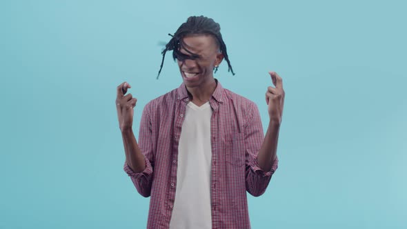 Black Man with Dreadlocks Crosses Fingers and Closing Eyes on a Blue Studio Background