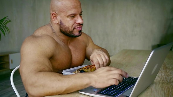 Powerful Man with Naked Chest Is Sitting and Typing on Laptop, Taking Fried Leg and Bitting It