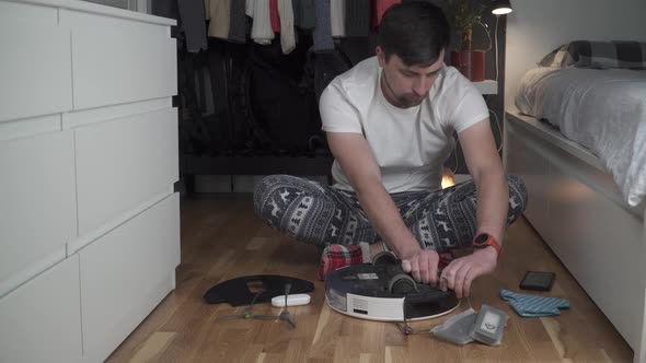 Caucasian Young Man in Christmas Pajamas Doing Vacuum Cleaner Robot Maintenance