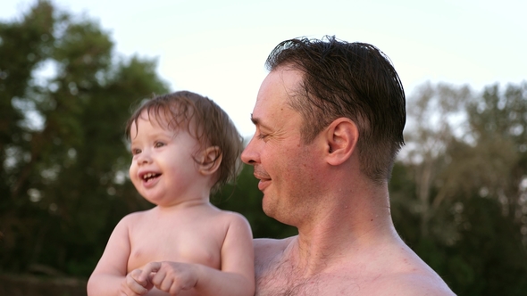 Small Wet Bathing Child Claps His Hands and Laughs Sitting in Embrace of Dad