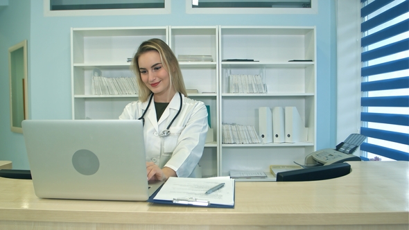 Smiling Medical Nurse Working on Laptop and Making Notes at Reception Desk