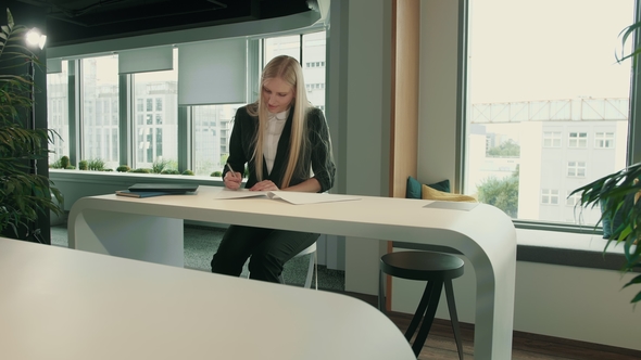 Elegant Woman Working in Stylish Office. Modern Blond Woman in Trendy Suit Sitting at Table in Light