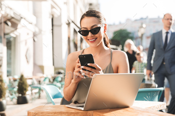glasses and dress holding cell phone while sitting in cozy cafe or restaurant outside in summer