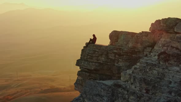 Aeril View a Man Sits on the Edge of a Cliff Against the Backdrop of a Mountain Panorama and Sunset