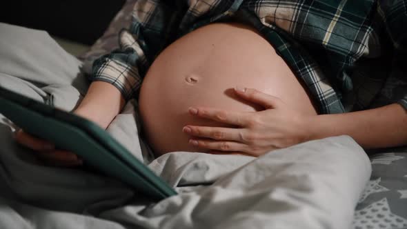 Pregnant with tablet computer in bedroom showing and touching belly.