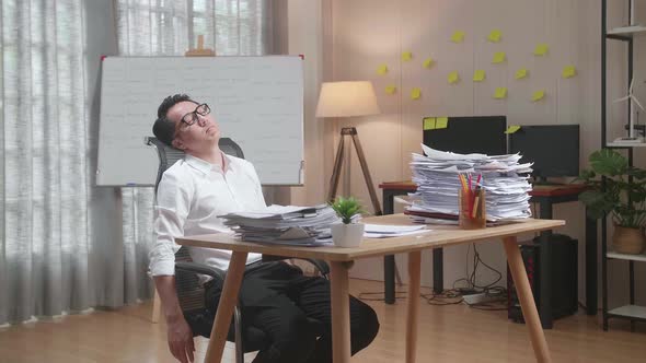 Tired Asian Man Leaning On The Office Chair And Sleeping While Working Hard With Documents