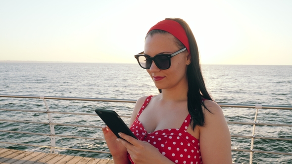 Pretty Woman Using Black Mobile Phone By the Sea Beach. Girl in Retro Red Polka Dots Dress. Sun