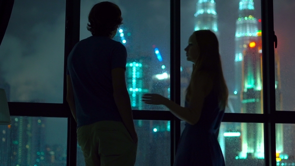 Silhouette of a Man and Woman Standing By a Panoramic Window at Night Kissing, Hugging and Looking