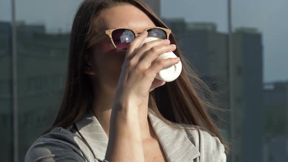 Smiling Young Woman in Sunglasses Drinks Coffee From a Disposable Cup