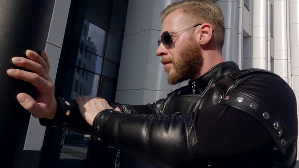 Stylish Young Man with a Beard in Sunglasses Stands Leaning His Hand on a Pillar in Fashionable