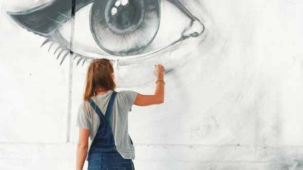 Beautiful Young Blonde Girl Making Graffiti of Big Eye with Aerosol Spray on Urban Street Wall