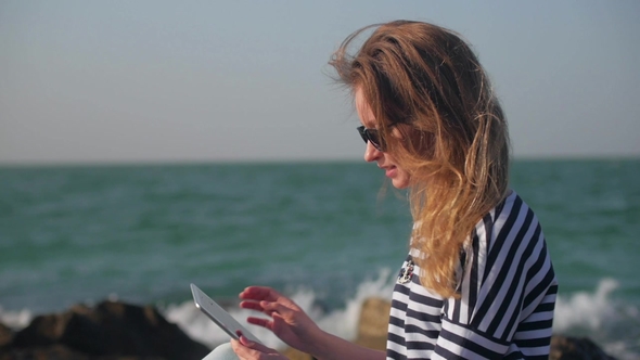 Cheerful Blonde Girl Is Watching Funny Photos in Her Tablet and Smiling Sitting Outdoors