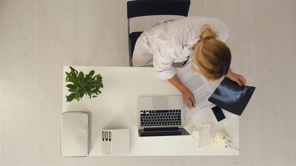 Young Female Doctor Working in the Office Holding X-ray Results and Using Her Laptop Computer