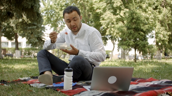 Young Businessman Sitting in Grass and Having Lunch in a Park Summertime