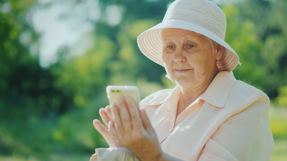An Elderly Woman in Summer Clothes and a Hat Uses a Smartphone. Relax in the Summer Park