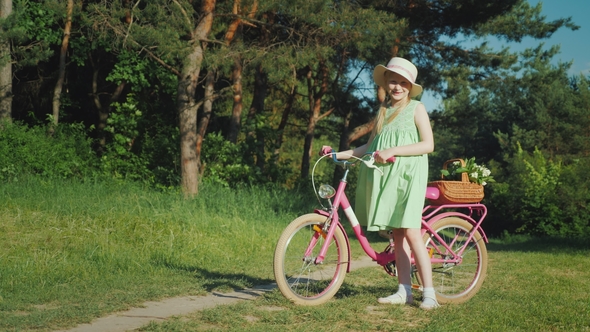 A Girl in a Summer Dress Is Standing Near a Bicycle, Looking at the Camera. With Her Basket of