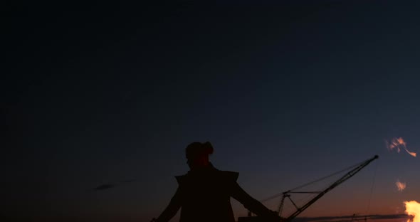 A Man in a Raincoat with Two Flamethrowers Lets Out a Fiery Flame Standing at Sunset on the Sand