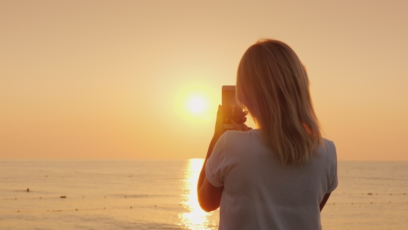 A Young Blonde Woman Is Photographing on a Smartphone a Pink Dawn By the Sea. View From the Back