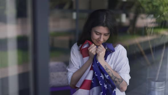 Medium Shot Satisfied Young Woman with American Flag Standing Indoors Looking Out the Window Smiling