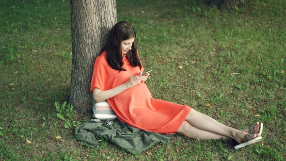 Relaxed Pregnant Girl Is Using Smartphone Touching Screen Sitting on Grass in Park on Warm Summer