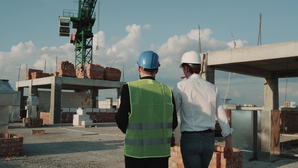 Two Young Engineers Walking Through Construction Site, Background Big Crane