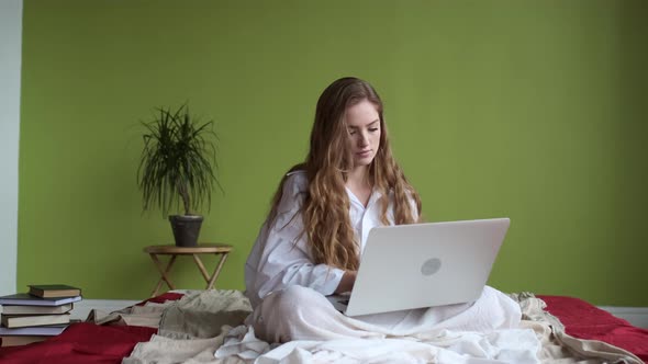 Young Girl Uses Laptop While Sitting On Bed In Apartment.