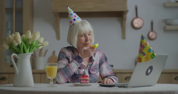 Cheerful Senior Woman Celebrates Birthday Online with Laptop, 70s Woman in Birthday Hat Blowing