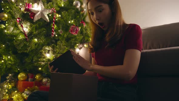 Young Asia female having fun opening X'Mas present box near Christmas tree decorated.